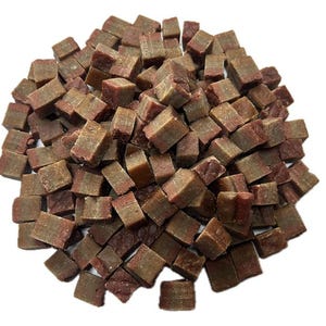 May include: A pile of small, square-shaped dog treats. The treats are a mix of brown and reddish-brown colors, with a layered appearance. The image is taken from a top-down perspective, showing the treats scattered across a white background.