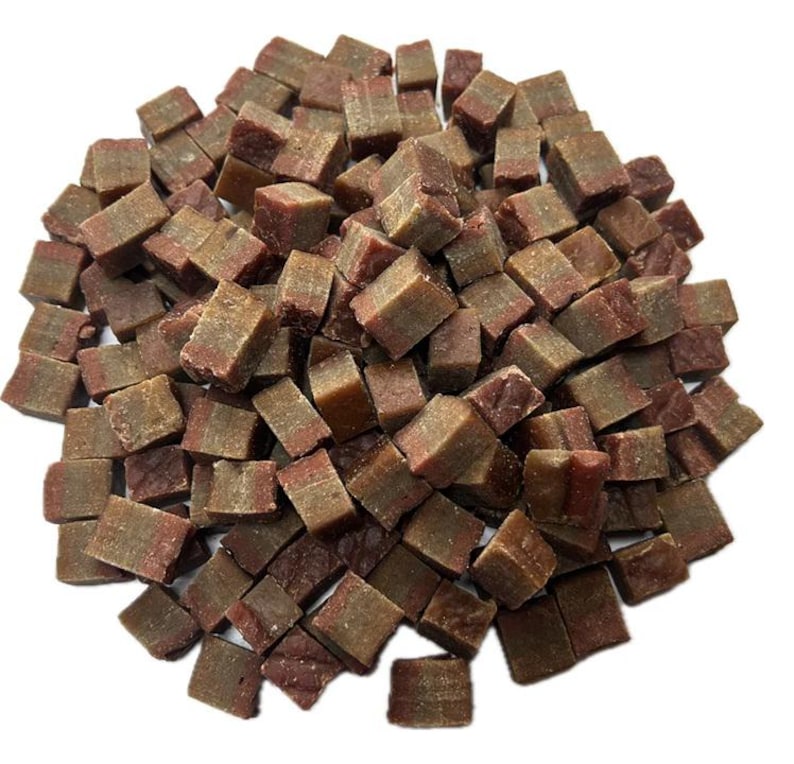 May include: A pile of small, square-shaped dog treats. The treats are a mix of brown and reddish-brown colors, with a layered appearance. The image is taken from a top-down perspective, showing the treats scattered across a white background.