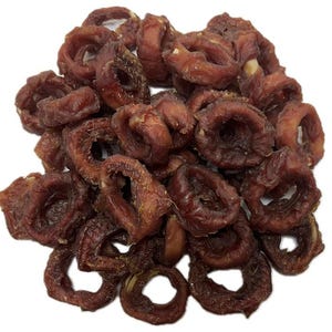 May include: A pile of dried, reddish-brown, ring-shaped treats, possibly pet food or snacks, against a white background. The treats have a slightly wrinkled texture and a dark, rich color, suggesting they are savory and possibly chewy.