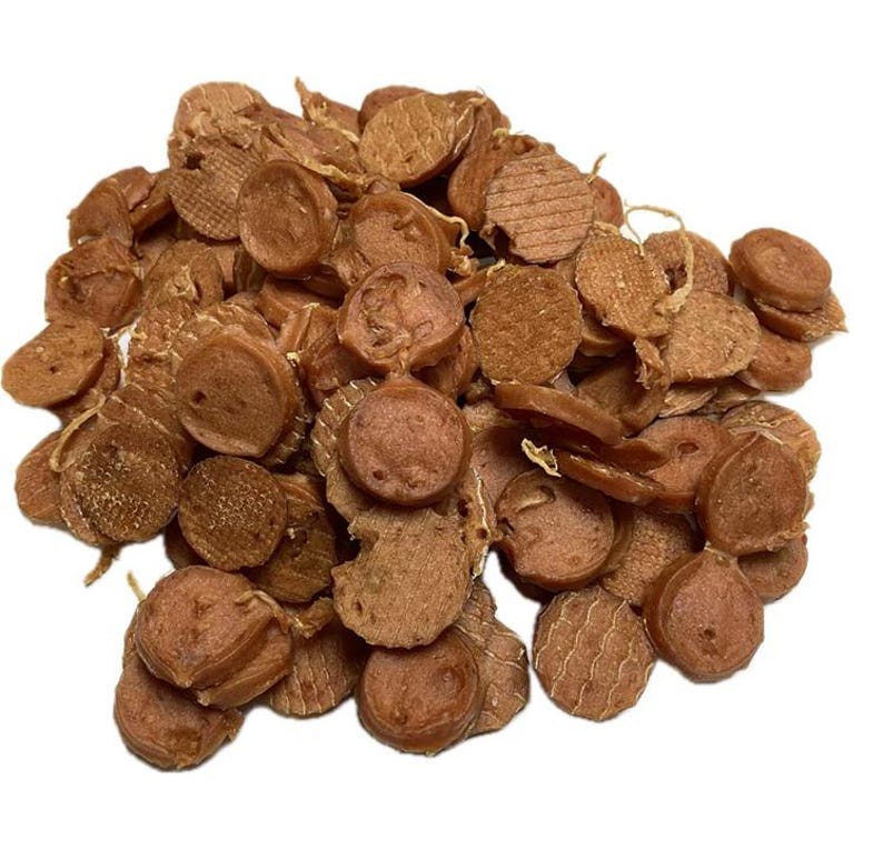 May include: A pile of brown, circular dog treats with various textures. Some treats are round, others are flat, and some have grill marks. The treats are scattered on a white background.