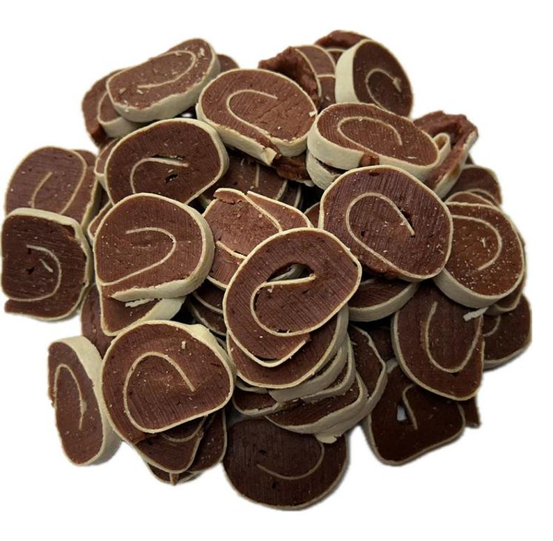 May include: A pile of dog treats, each a dark brown color with a white outer edge and a light-colored swirl in the center. The treats are arranged in a random pattern against a white background.