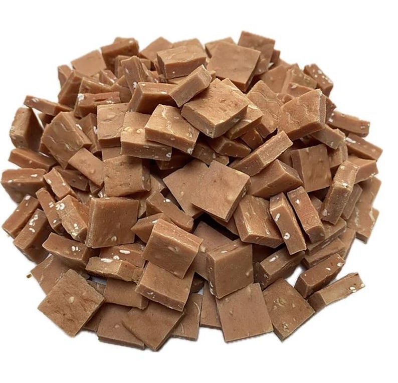 May include: A pile of small, square-shaped pieces of fudge. The fudge is a light brown color with visible white specks throughout. The pieces are scattered, creating a textured and appetizing appearance.