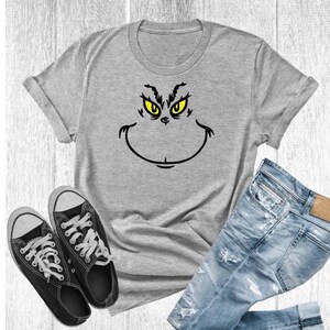 Pu&ograve; includere: Una maglietta grigia con una grafica del viso del Grinch in nero e giallo. La maglietta &egrave; mostrata su uno sfondo di legno bianco con un paio di scarpe da ginnastica nere e jeans blu.