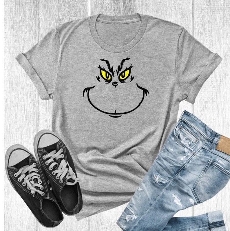 Pu&ograve; includere: Una maglietta grigia con una grafica del viso del Grinch in nero e giallo. La maglietta &egrave; mostrata su uno sfondo di legno bianco con un paio di scarpe da ginnastica nere e jeans blu.