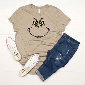 Pu&ograve; includere: Una maglietta color beige con una grafica del viso del Grinch in nero e giallo. La maglietta &egrave; abbinata a jeans blu e scarpe da ginnastica bianche.