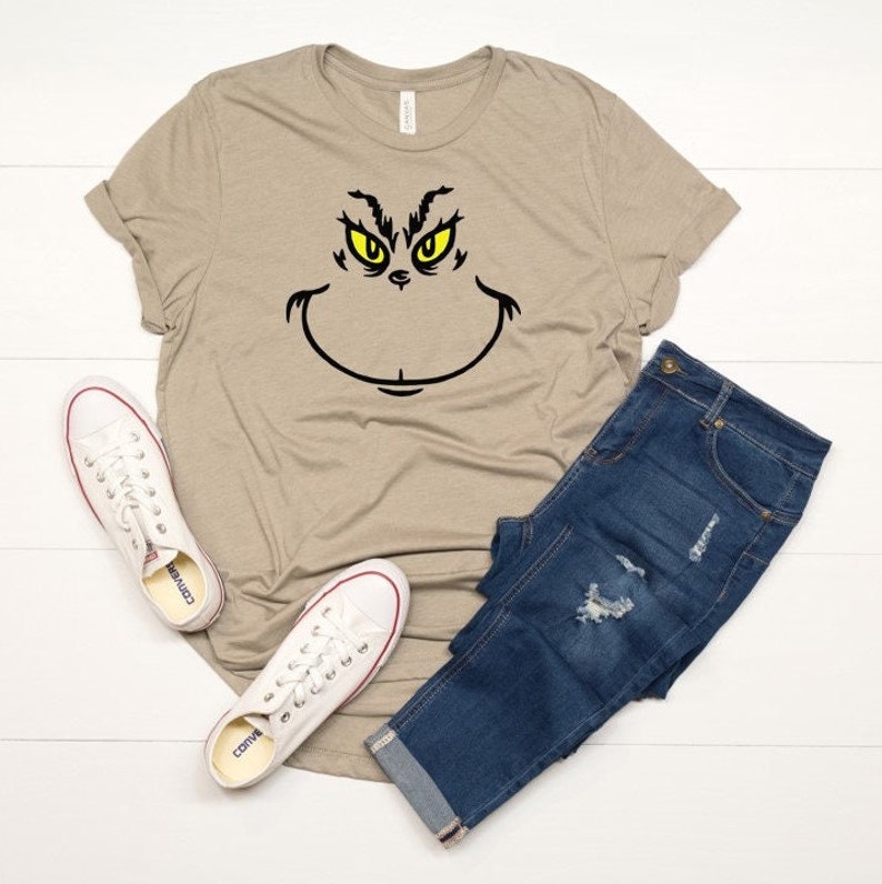 Pu&ograve; includere: Una maglietta color beige con una grafica del viso del Grinch in nero e giallo. La maglietta &egrave; abbinata a jeans blu e scarpe da ginnastica bianche.
