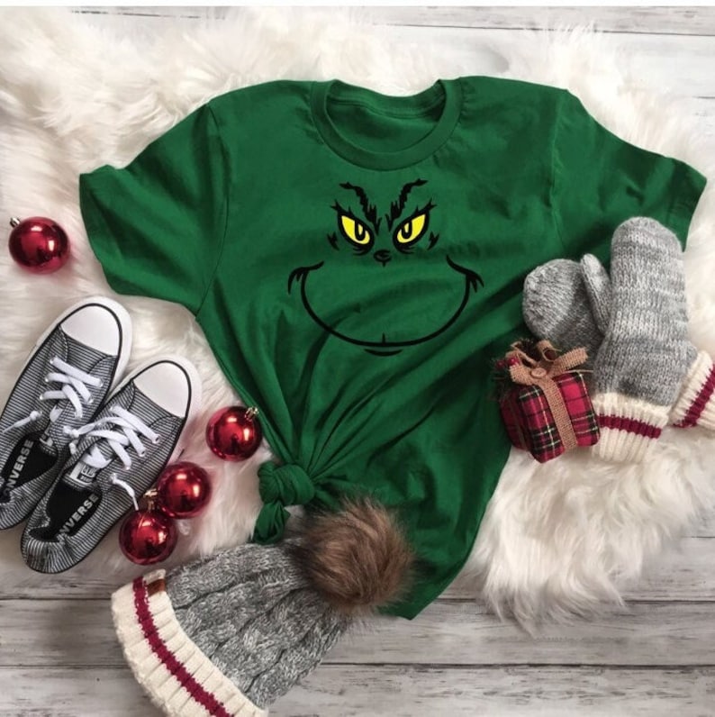 Pu&ograve; includere: Maglietta verde con una grafica del viso del Grinch. Gli occhi del Grinch sono gialli e la bocca &egrave; nera. La maglietta &egrave; su una superficie bianca e soffice con ornamenti rossi e bianchi, una piccola scatola regalo e un paio di guanti grigi.