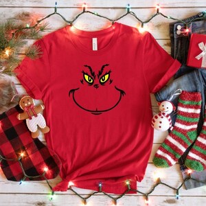 Pu&ograve; includere: Maglietta rossa con una grafica del viso del Grinch in nero e giallo. Il Grinch sta sorridendo con gli occhi spalancati.