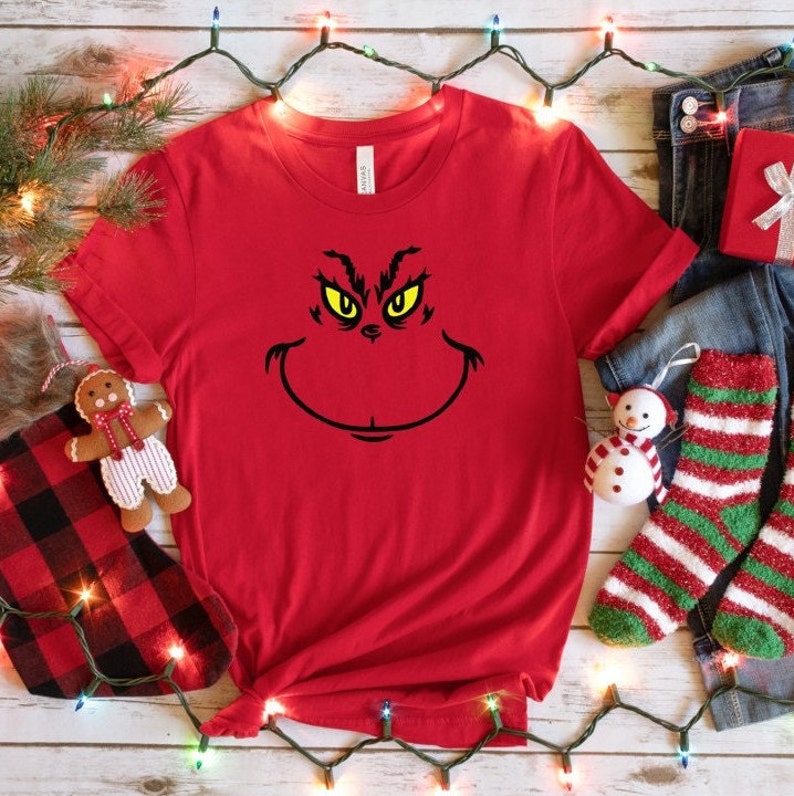 Pu&ograve; includere: Maglietta rossa con una grafica del viso del Grinch in nero e giallo. Il Grinch sta sorridendo con gli occhi spalancati.