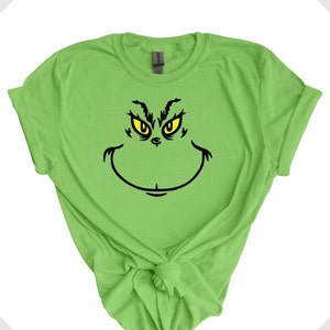 Pu&ograve; includere: Una maglietta verde con una grafica del volto del Grinch in nero e giallo.