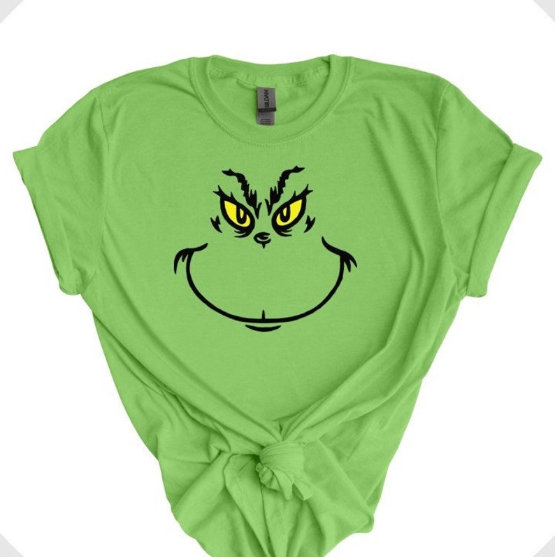 Pu&ograve; includere: Una maglietta verde con una grafica del volto del Grinch in nero e giallo.