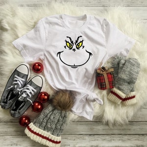 Pu&ograve; includere: Maglietta bianca con una grafica del viso del Grinch in nero e giallo. La maglietta &egrave; su una superficie bianca e soffice con ornamenti rossi e grigi, un berretto grigio con un pon pon marrone e guanti grigi.