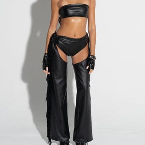 May include: A black two-piece outfit, comprising a strapless top and high-waisted bottoms. The ensemble includes matching chaps with fringe detailing and fingerless gloves. The outfit is made of a shiny, textured material.
