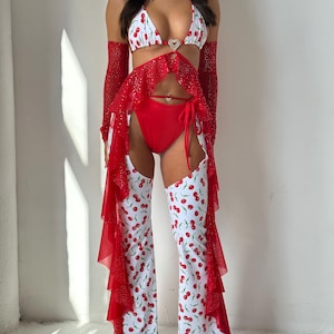 Puede incluir: Conjunto de bikini y pantalón blancos con estampado de cerezas rojas. El conjunto incluye una braguita de bikini roja, mangas rojas y una falda con volantes roja. Una gema en forma de corazón adorna la parte superior del bikini.