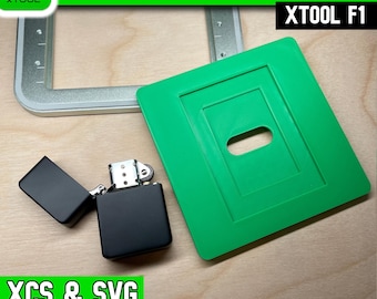 May include: A green plastic flip top lighter jig for the xTool F1 laser machine, featuring a rectangular cutout. A matte black flip top lighter is positioned next to the jig, and a partial silver metal frame is visible in the background.