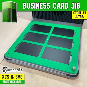 May include: A green business card jig for the xTool F1 Ultra laser machine. The rectangular frame features six rectangular cutouts for holding business cards in place. The device includes silver metal knobs and is labeled with the text Samcraft.