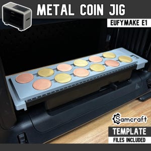 May include: A gray metal coin jig for the EufyMake E1, featuring a rectangular tray with twelve circular indentations holding alternating gold and copper-colored metal discs. The jig includes a printed ruler along the front edge and text.
