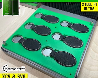 Op de afbeelding: Een felgroene sleutelhanger-mal ontworpen voor de xTool F1 Ultra, met zes ovale uitsparingen die zwarte kunstlederen sleutelhangers met zilveren metalen inzetstukken bevatten. De mal bevat metalen uitlijnpinnen en is gelabeld met de tekst KEYCHAIN JIG, XTOOL F1 ULTRA en Samcraft XCS & SVG FILES INCLUDED.
