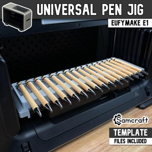Puede incluir: Una plantilla universal gris para bolígrafos para el grabador láser EufyMake E1, que sostiene doce bolígrafos de madera clara en ranuras paralelas. El dispositivo está dentro de una máquina negra, con los textos Universal Pen Jig y Samcraft.