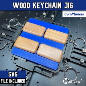 May include: A black plastic keychain jig with a blue insert holding four rectangular wooden keychain blanks. The jig features adjustable metal screws and is attached to metal key rings with chains. The text WOOD KEYCHAIN JIG, ComMarker, and Samcraft are visible.
