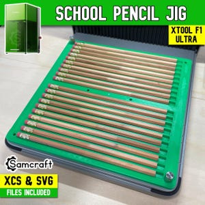 May include: A green plastic school pencil jig designed for the xTool F1 Ultra, featuring multiple parallel slots holding wooden pencils with metal ferrules. The device includes text labels reading SCHOOL PENCIL JIG, XTOOL F1 ULTRA, and Samcraft XCS & SVG FILES INCLUDED.