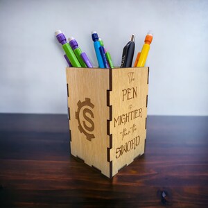 May include: Wooden pencil holder with the quote "The pen is mightier than the sword" engraved on the side. The holder is filled with pens and pencils in various colors.