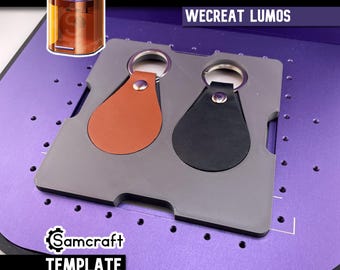Op de afbeelding: Twee druppelvormige sleutelhangers, een in lichtbruin en een in zwart, zijn bevestigd met zilverkleurige metalen ringen en klinknagels. Ze liggen in een grijze rechthoekige sjabloonhouder met uitsparingen, gelabeld Teardrop Keychain Wecreat Lumos.