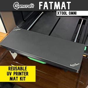 May include: A black Samcraft Fatmat reusable UV printer mat kit for the xTool Omni. The rectangular mat features a smooth surface with the Samcraft Fatmat logo printed in white on the corners and a bright green xTool accent piece attached.