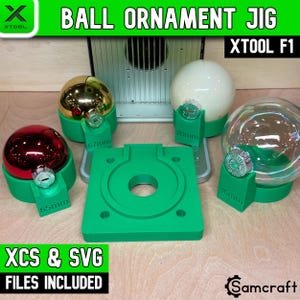 May include: A green plastic ball ornament jig for the xTool F1 laser machine, featuring a square base and four circular holders labeled 65mm, 67mm, 80mm, and 86mm. The set includes various colored spherical ornaments resting in the holders.