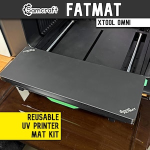 May include: A black Samcraft Fatmat reusable UV printer mat kit for the xTool Omni. The rectangular mat features the Samcraft Fatmat logo in white on the top left and bottom right corners, with a bright green xTool accent piece visible underneath.