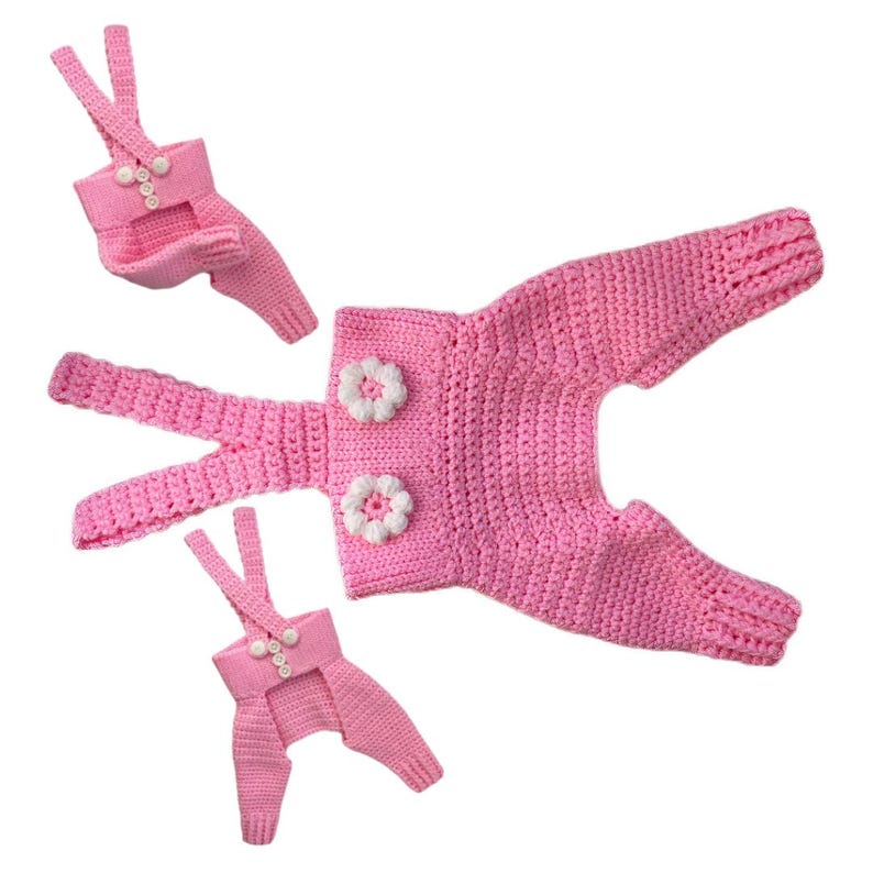 May include: Three pink crocheted overalls of varying sizes, each featuring thin shoulder straps and white buttons. The largest pair is adorned with two white crocheted flower embellishments on the front bodice, while the smaller pairs feature three vertical white buttons on the chest.