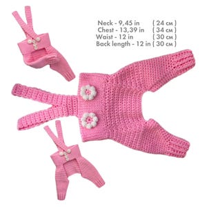 May include: Three pink crocheted overalls with white flower accents and straps. Text: Neck - 9.45 in, Chest - 13.39 in, Waist - 12 in, Back length - 12 in. The garments feature a textured knit pattern and button closures on the straps.