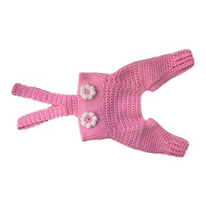May include: A light pink crochet overall garment featuring two thin shoulder straps and two white crocheted flower embellishments attached to the front bodice. The piece is constructed with a textured yarn stitch pattern throughout.
