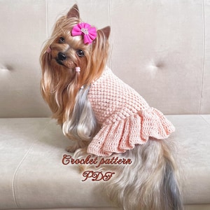 May include: A small, light pink crocheted dog dress with a ruffled hem. The dress is being worn by a Yorkshire Terrier with a pink bow in its hair. The text "Crochet pattern PDF" is written below the dog.