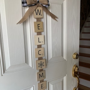 May include: A welcome sign made of wooden tiles with the letters "WELCOME" spelled out. The tiles are strung together with a burlap ribbon and a black and white checkered bow. A snowflake tile is included in the sign.
