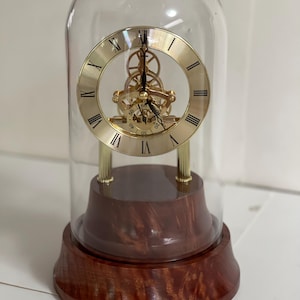 May include: A gold-toned clock with exposed gears is encased in a glass dome on a wooden base. The clock face has Roman numerals and a black hour and minute hand.