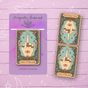 May include: A set of magnetic bookmarks with a tarot card design. The cards feature a hand with the middle finger extended, surrounded by floral elements and an eye. The text "THE FINGER" is displayed on the cards.