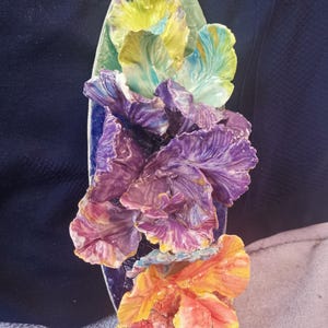 Puede incluir: Una escultura de pared de cerámica con tres flores estilizadas en tonos amarillos, morados y naranjas. Las flores están dispuestas verticalmente sobre un fondo ovalado azul y verde.