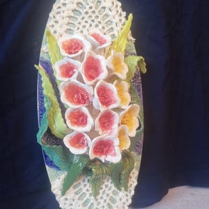 Puede incluir: Una decoración de pared de cerámica con un ramo de flores blancas y rojas con detalles amarillos. Las flores están dispuestas sobre un fondo ovalado blanco con un diseño de encaje.