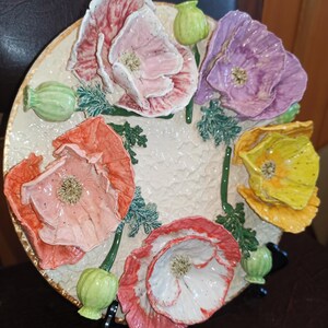 Puede incluir: Un plato de cerámica decorativo con cinco flores de amapola esculpidas en rosa, morado, naranja y amarillo. El plato tiene un fondo blanco texturizado y un borde dorado.
