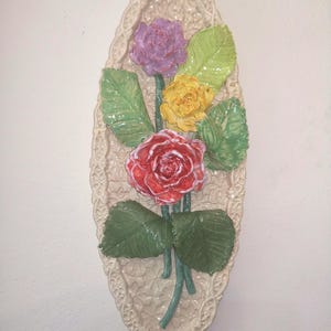 Puede incluir: Un adorno de pared de cerámica con un ramo de rosas rojas, moradas y amarillas, con hojas verdes. Las rosas están dispuestas sobre una base ovalada color crema con un diseño de encaje.