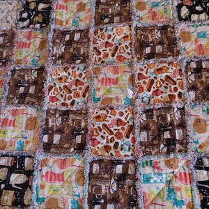 May include: A patchwork quilt with a variety of brown, white, and blue fabric squares. Each square features a different pattern, including coffee cups, pastries, and other food items. The quilt is made with a frayed edge, giving it a rustic look.