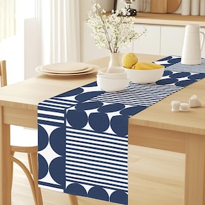 Puede incluir: Un camino de mesa geométrico azul y blanco con un patrón de círculos y rayas. El camino de mesa está sobre una mesa de madera con un jarrón de flores blancas y un cuenco de limones.