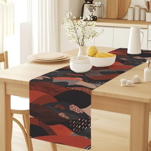May include: A black, red and white patterned table runner with abstract shapes and dots. The runner is on a wooden table with a vase of white flowers and a bowl of lemons.