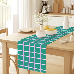 Pode incluir: Um corredor de mesa xadrez verde e rosa em uma mesa de madeira com uma tigela de frutas e um vaso de flores brancas.