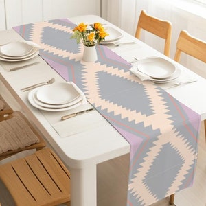 May include: A dining table set for a meal, featuring a patterned table runner. The runner has a geometric design in gray, peach, and lavender. White plates, silverware, and a vase of yellow flowers complete the setting. Wooden chairs surround the table.