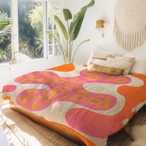 Op de afbeelding: Een beige dekbedovertrek met een abstract patroon in roze, oranje en geel. Het dekbedovertrek ligt op een bed met witte kussens en een bruin kussen.