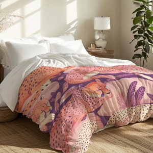 May include: A pink and orange throw blanket with a cheetah print pattern. The blanket is on a bed with white bedding and a wooden nightstand.
