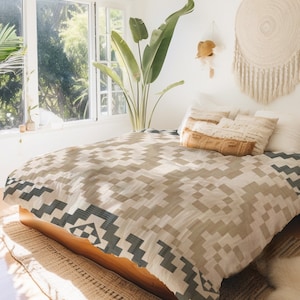 Puede incluir: Una manta de tiro de color beige y gris con un diseño geométrico. La manta está sobre una cama con almohadas blancas y un marco de madera.