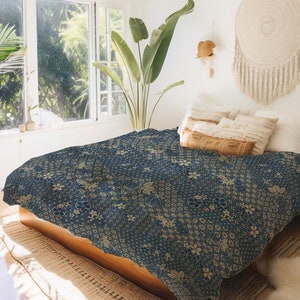 May include: A blue and gold patterned duvet cover with a floral design. The duvet cover is on a bed with white pillows and a brown throw pillow. The bed is in a bedroom with a window and a plant.